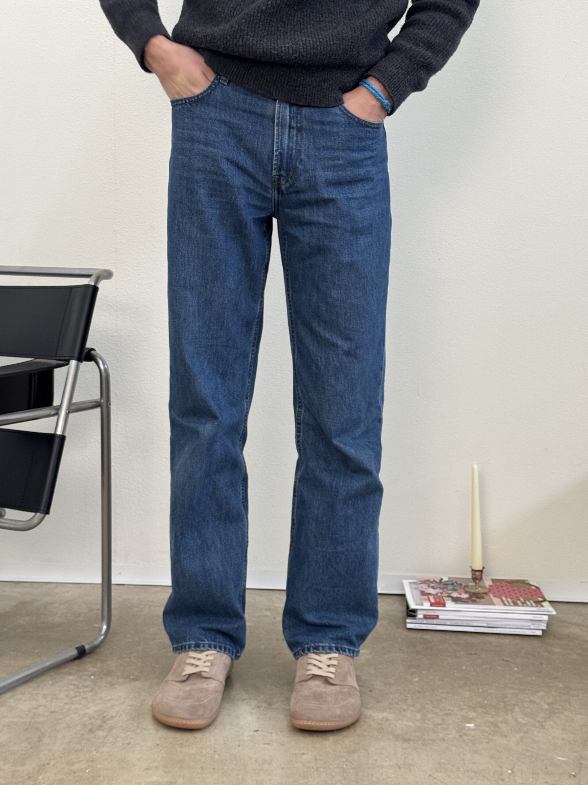 A person wearing straight-leg blue denim jeans and beige suede sneakers, standing with hands in pockets. In the background, a minimalist interior with a black chair and a stack of magazines on the floor.